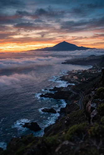 A high-angle view over a rugged coastline at twilight. The ocean churns against dark, rocky shores while a winding road threads through the settlements below, and a blanket of low cloud stretches out to sea in front of a distant mountain silhouette, under a sky streaked with orange and blue.