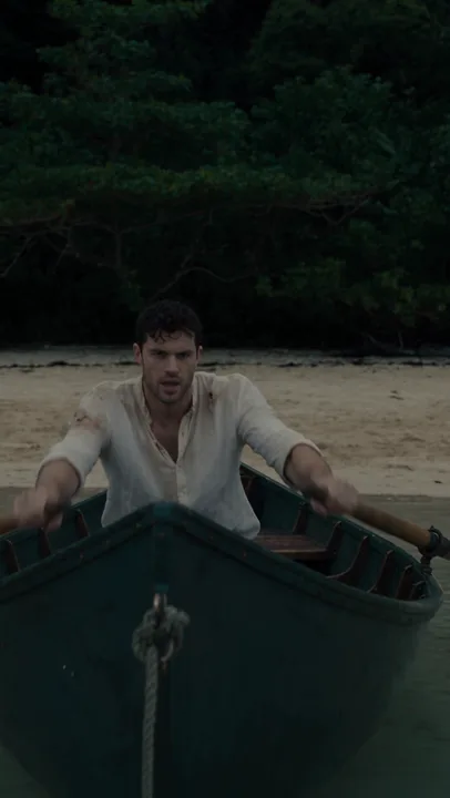 A man in a rowboat approaches a sandy shore next to rocky outcrops under a cloudy sky. He is exiting the boat, looking to his right with a startled expression. The low-angle shot and muted colors create a sense of unease and suspense. The lighting is dim, suggesting either dawn, dusk, or an overcast day.