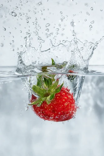 A vibrant red strawberry plunges into clear water, creating a dynamic splash with suspended droplets. The high-speed capture freezes the motion, highlighting the fruit's texture and the water's ephemeral nature. The lighting is bright and clean, emphasizing the subject against a softly blurred background.